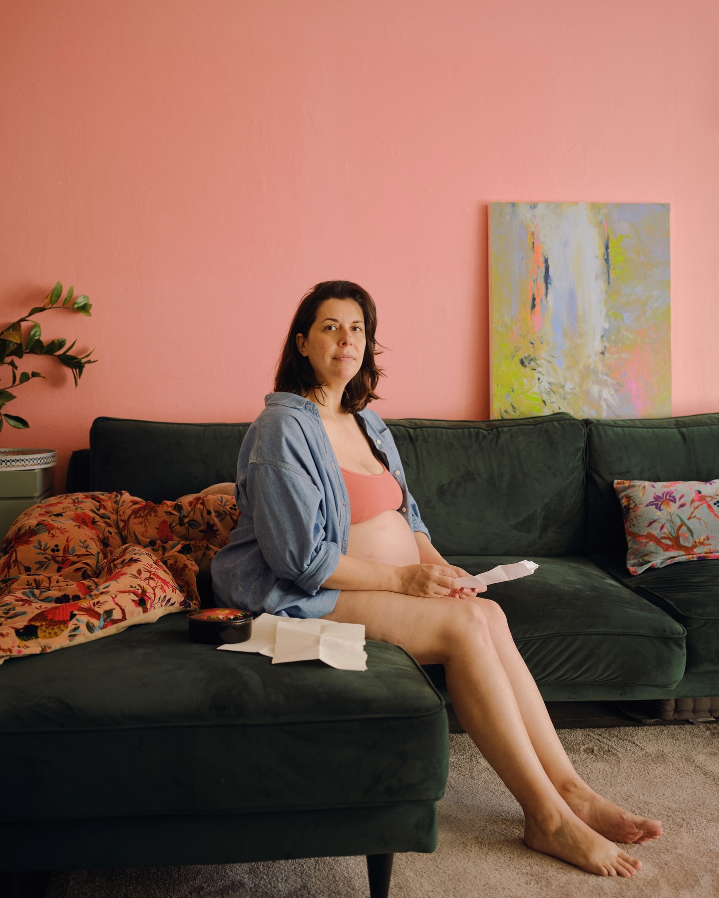 Pregnant woman sitting on a green sofa in a living room with pink walls and an abstract painting, wearing a blue shirt and peach bra.