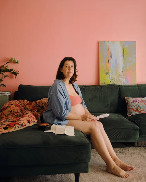 Pregnant woman sitting on a green sofa in a living room with pink walls and an abstract painting, wearing a blue shirt and peach bra.