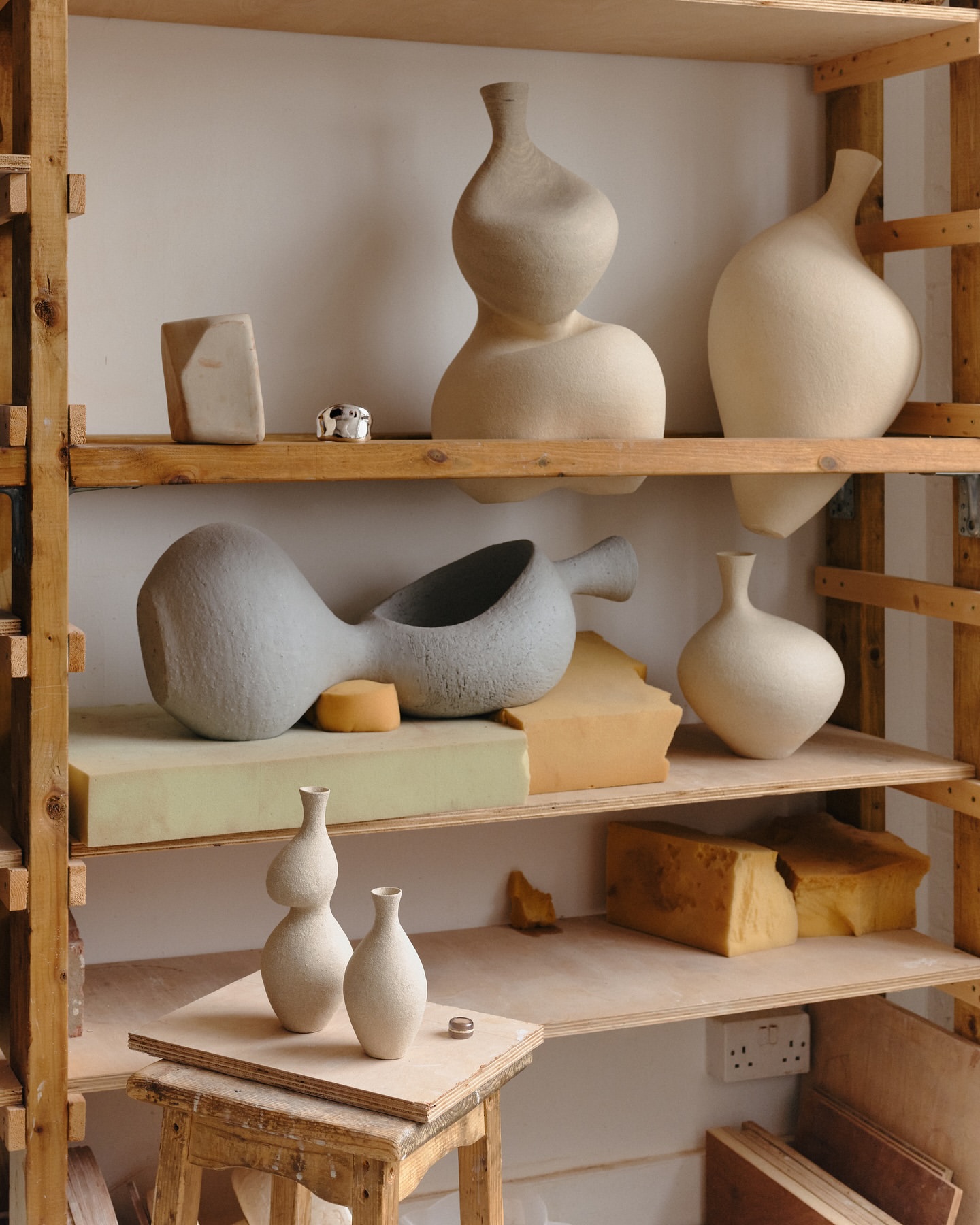 Still life composition featuring handmade ceramic vases and objects arranged on a wooden shelf in a minimalist style. Neutral color palette.