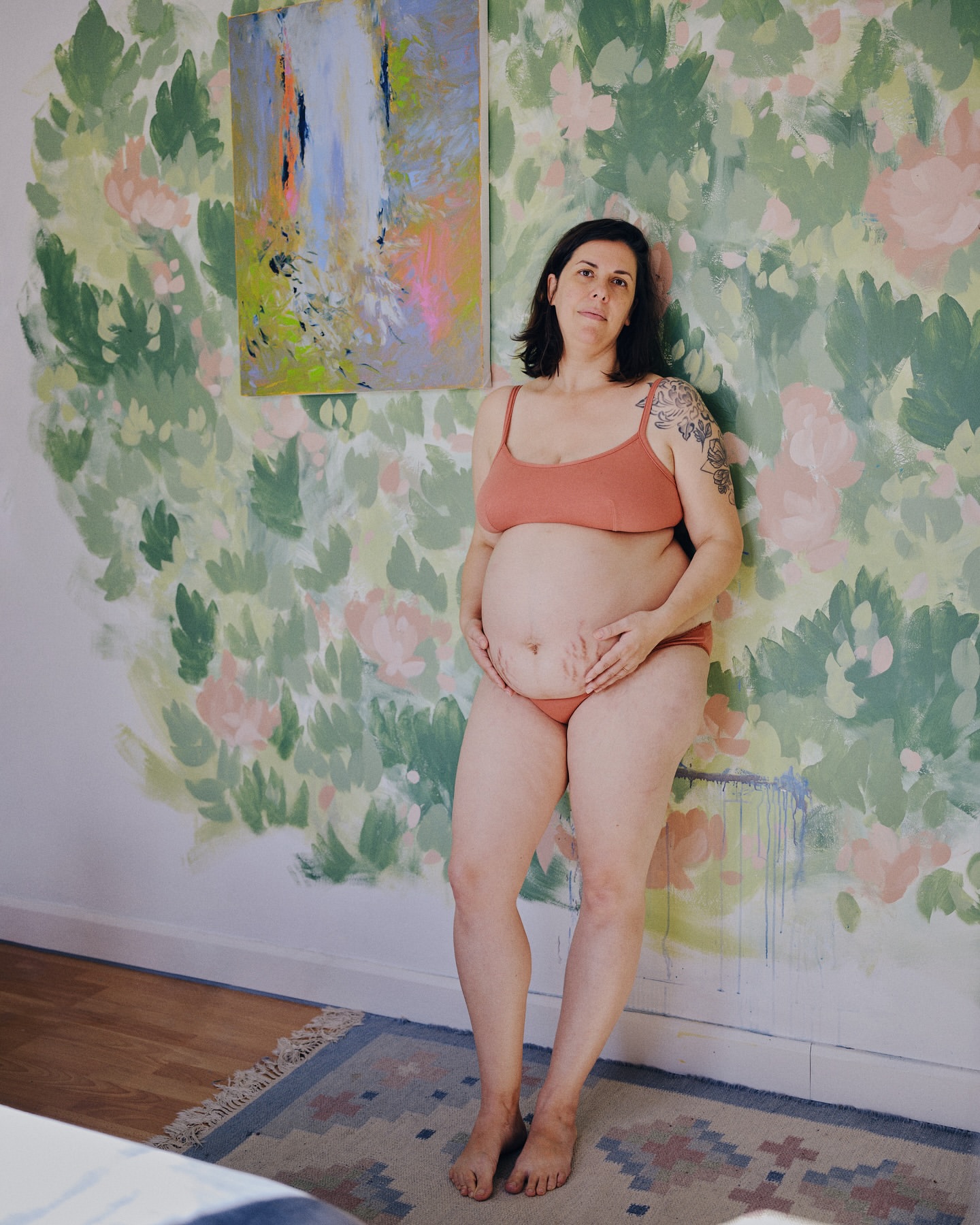 Pregnant woman in lingerie poses against a hand-painted floral wall with an abstract painting, shot in natural light indoors.