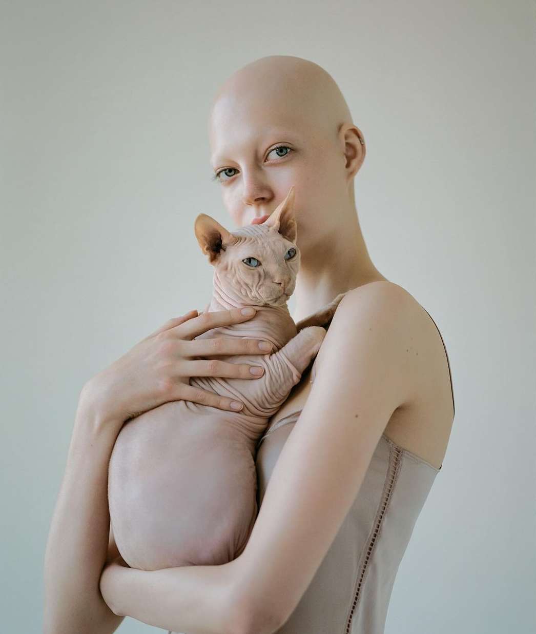 Close-up shot of a bald adult female gently holding a Sphynx cat, both in a light-beige palette against a pale-beige indoor backdrop.