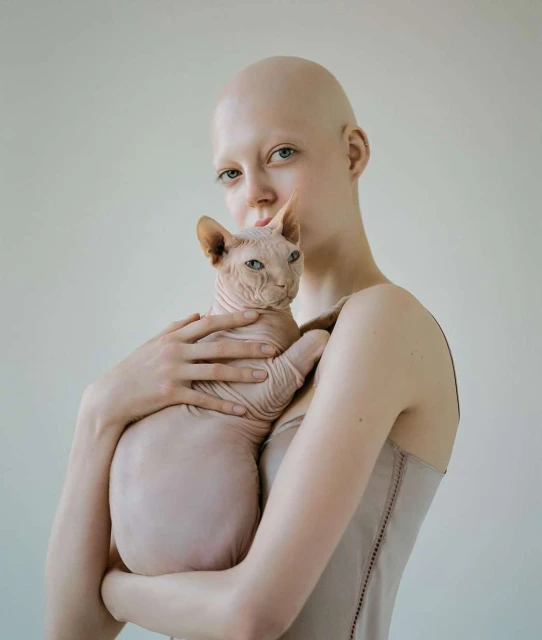 Close-up shot of a bald adult female gently holding a Sphynx cat, both in a light-beige palette against a pale-beige indoor backdrop.