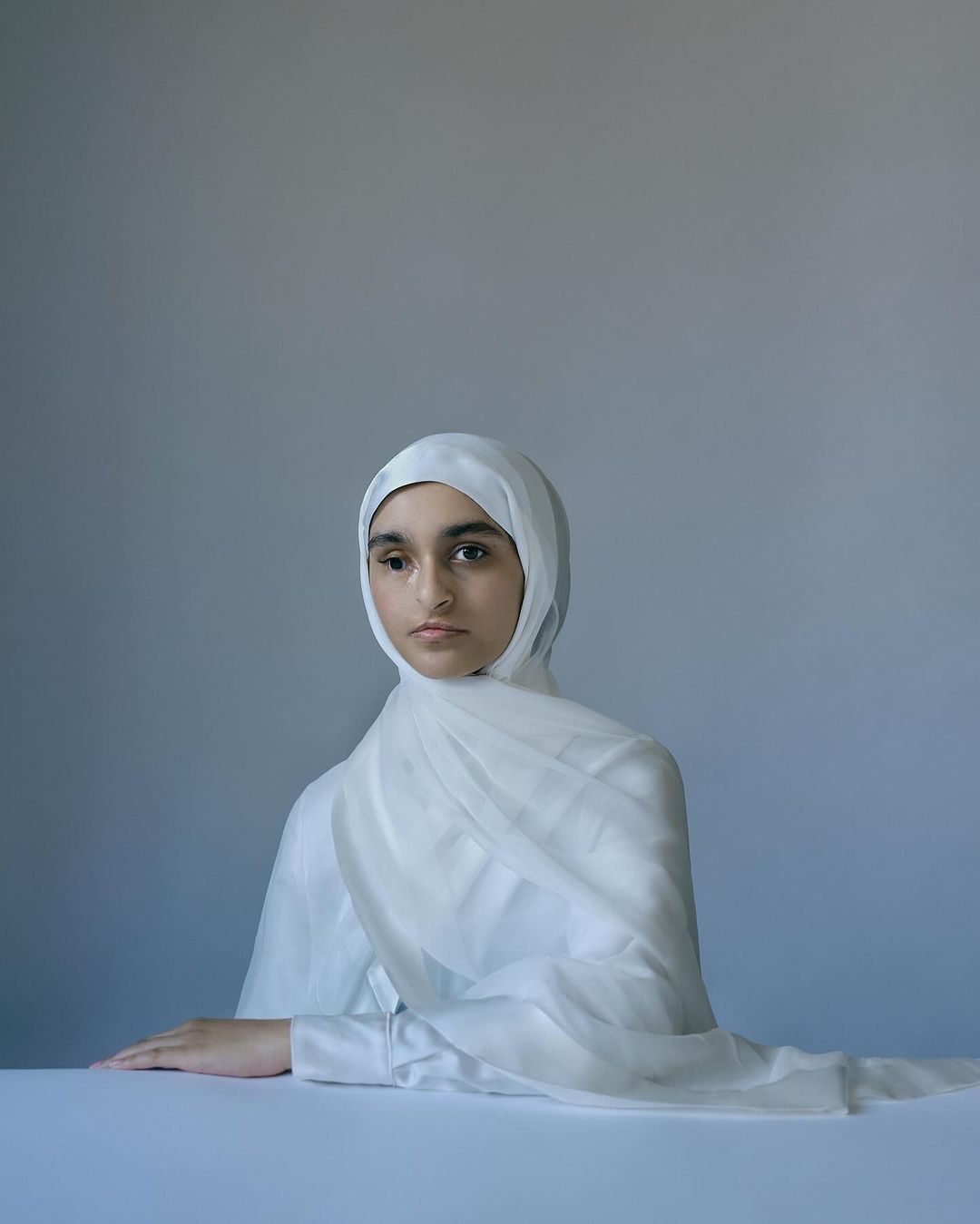 Portrait of a young woman in a white hijab and matching shirt, seated at a table against a light grey background in a studio setting.