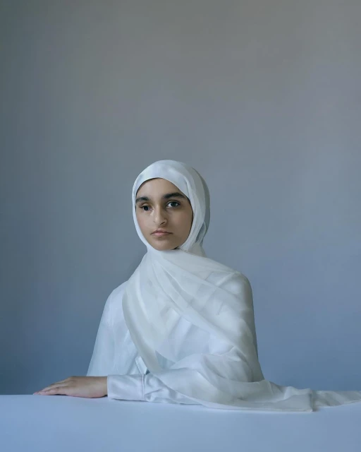 Portrait of a young woman in a white hijab and matching shirt, seated at a table against a light grey background in a studio setting.