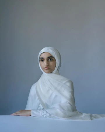 Portrait of a young woman in a white hijab and matching shirt, seated at a table against a light grey background in a studio setting.