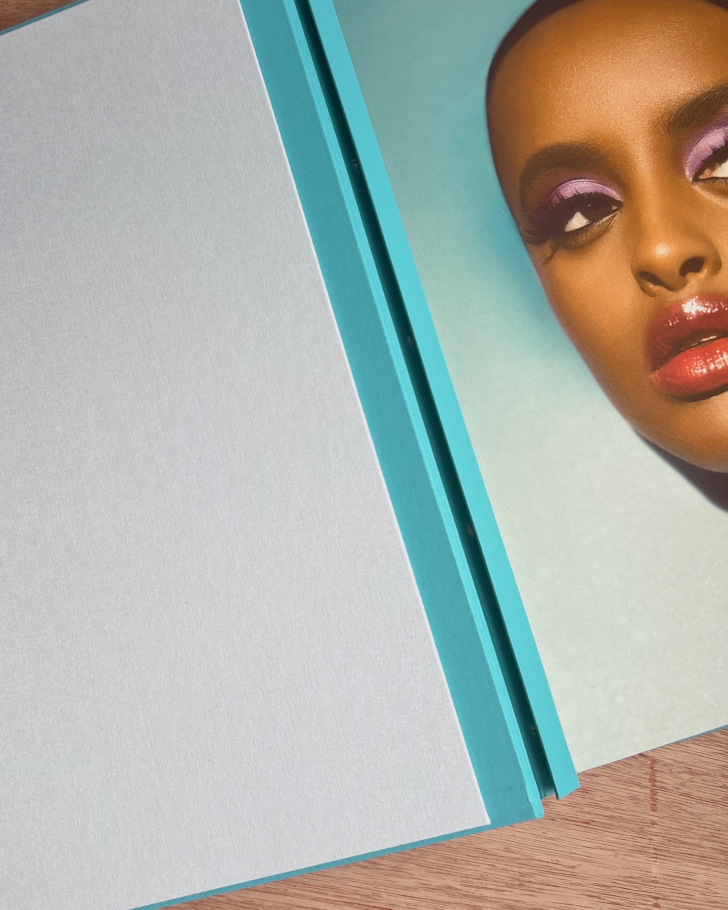Close-up of a dark-skinned woman with dramatic purple eyeshadow and glossy lips, alongside a light blue flat surface with a turquoise edge.