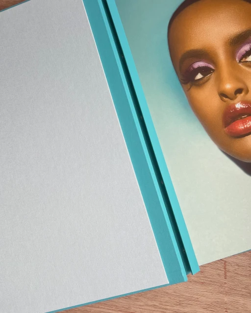 Close-up of a dark-skinned woman with dramatic purple eyeshadow and glossy lips, alongside a light blue flat surface with a turquoise edge.