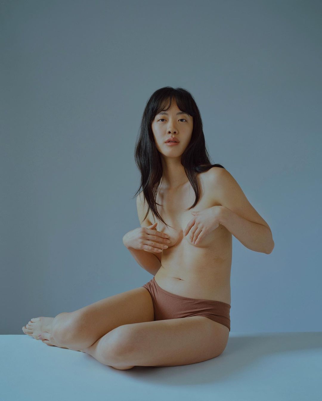 Portrait of an Asian adult female in underwear sitting cross-legged with hands over her chest against a blue and white background.