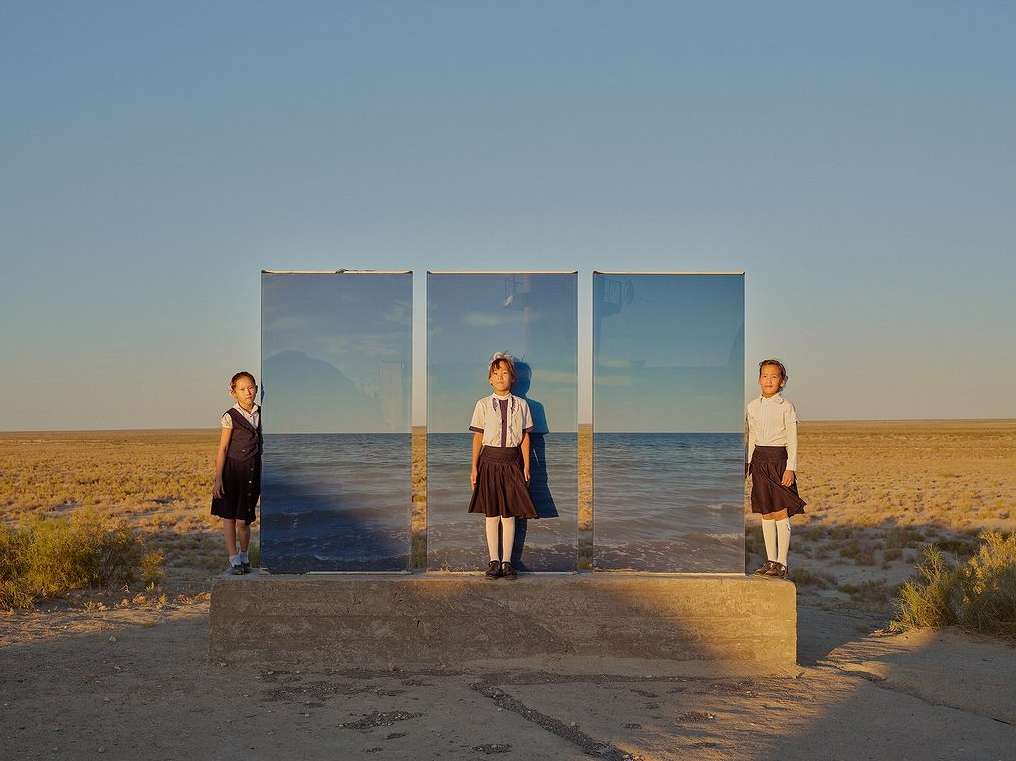 Three schoolgirls stand in front of three reflective panels in a tan field under a blue sky, mirroring the ocean.
