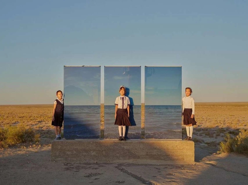 Three schoolgirls stand in front of three reflective panels in a tan field under a blue sky, mirroring the ocean.