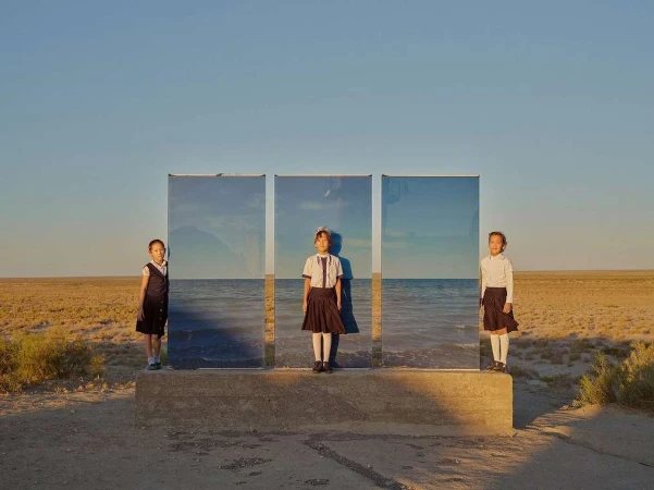 Three schoolgirls stand in front of three reflective panels in a tan field under a blue sky, mirroring the ocean.