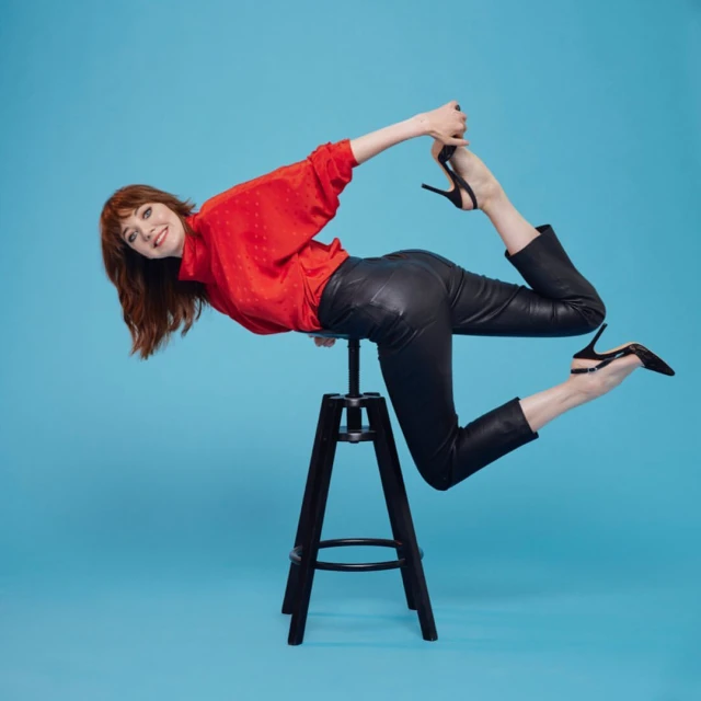 Woman in red blouse and black leather pants posing on a stool with black high heels against a blue backdrop in a studio setting.
