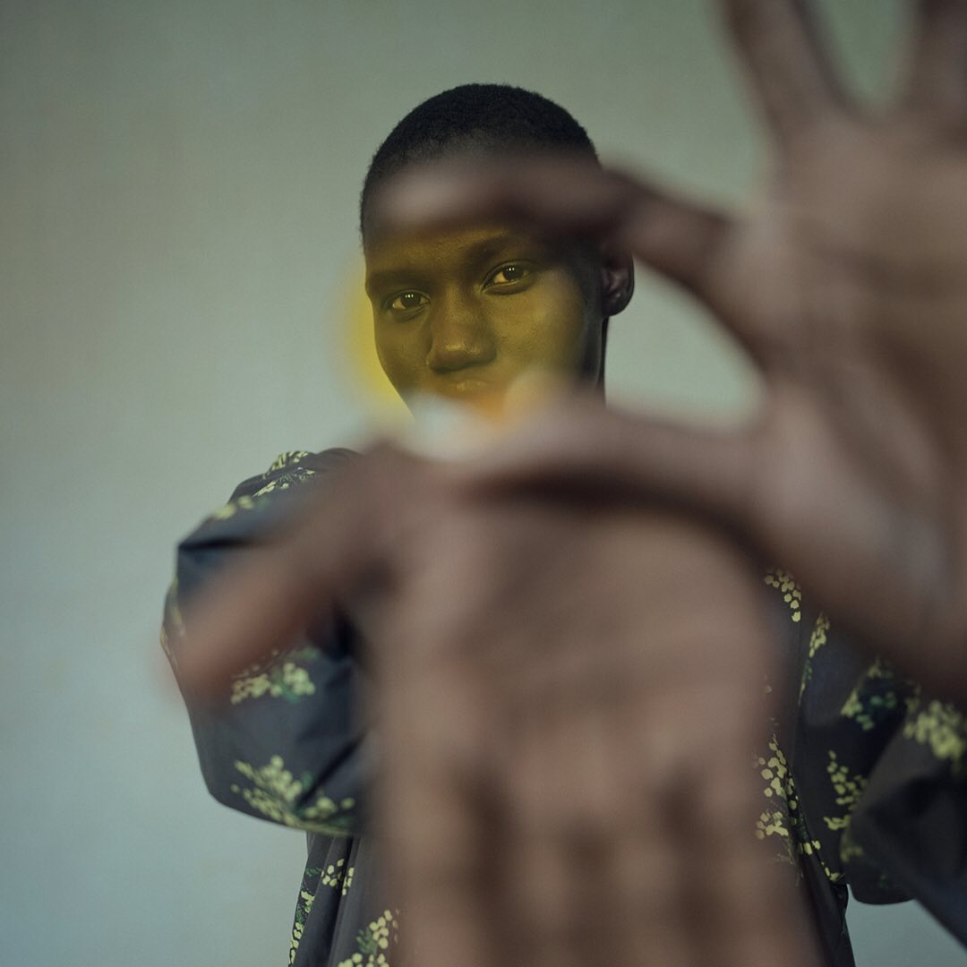 Portrait of a dark-skinned adult female with short hair wearing floral print top, holding hands out towards camera in an indoor setting.