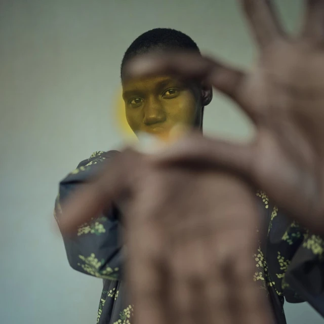 Portrait of a dark-skinned adult female with short hair wearing floral print top, holding hands out towards camera in an indoor setting.