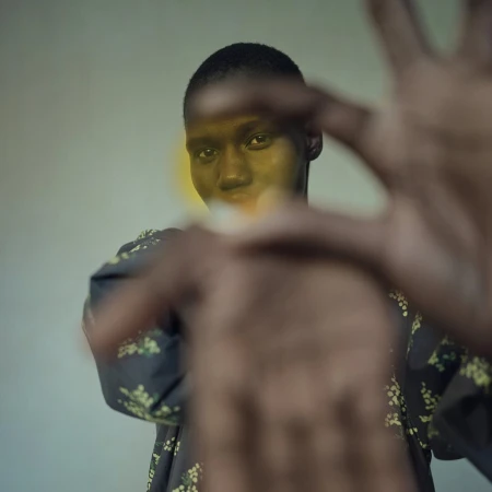 Portrait of a dark-skinned adult female with short hair wearing floral print top, holding hands out towards camera in an indoor setting.