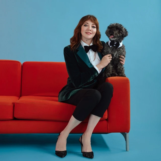 Adult woman with auburn hair sits on a red couch with a black dog wearing a bow tie against a blue backdrop in an indoor studio.