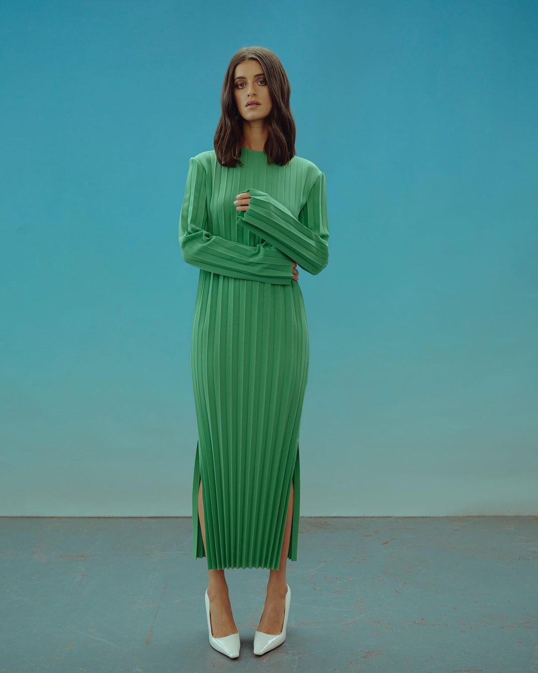 Elegant woman in a pleated green dress with long sleeves and white heels stands against a blue background in a full-length portrait.