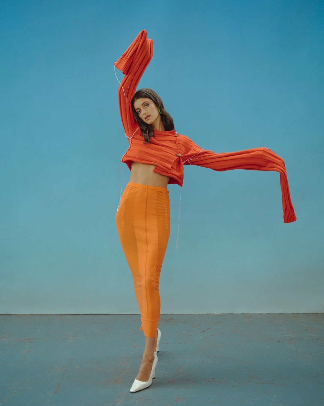 A woman in an orange top and skirt poses against a blue backdrop, her arms outstretched, creating a fashion editorial style image.