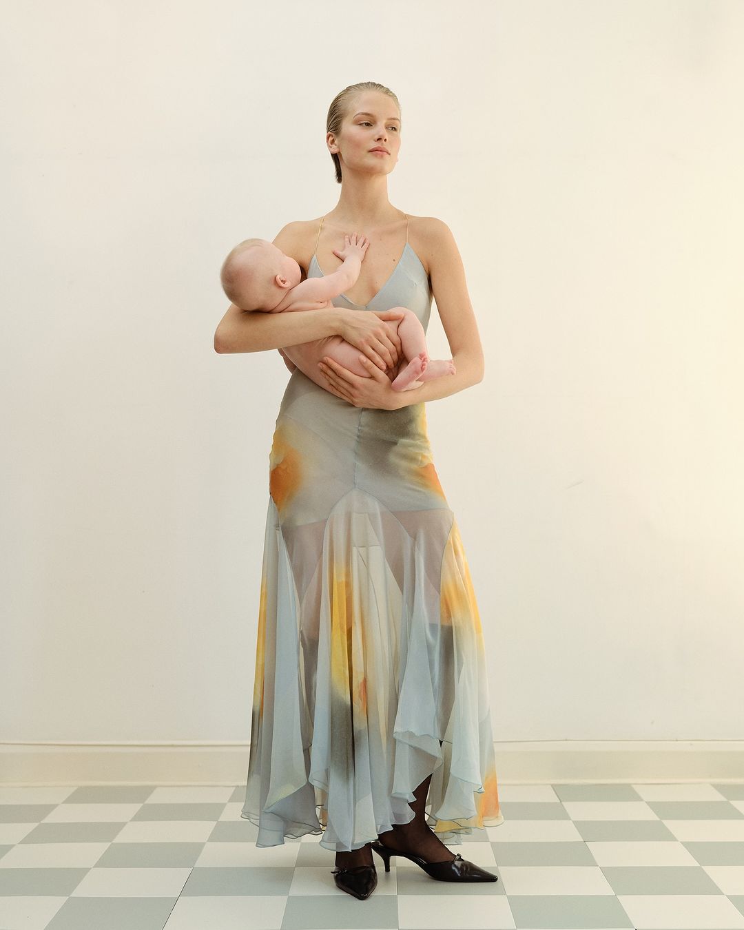 Adult female fashion model wearing a flowing dress, holding a baby in her arms in an indoor setting with a checkered floor.