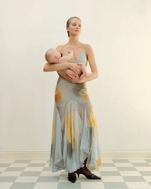 Adult female fashion model wearing a flowing dress, holding a baby in her arms in an indoor setting with a checkered floor.