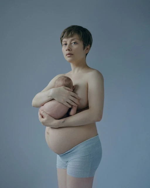 Portrait of a pregnant woman holding a newborn baby. The woman has short hair and is topless with blue underwear. Grey background.