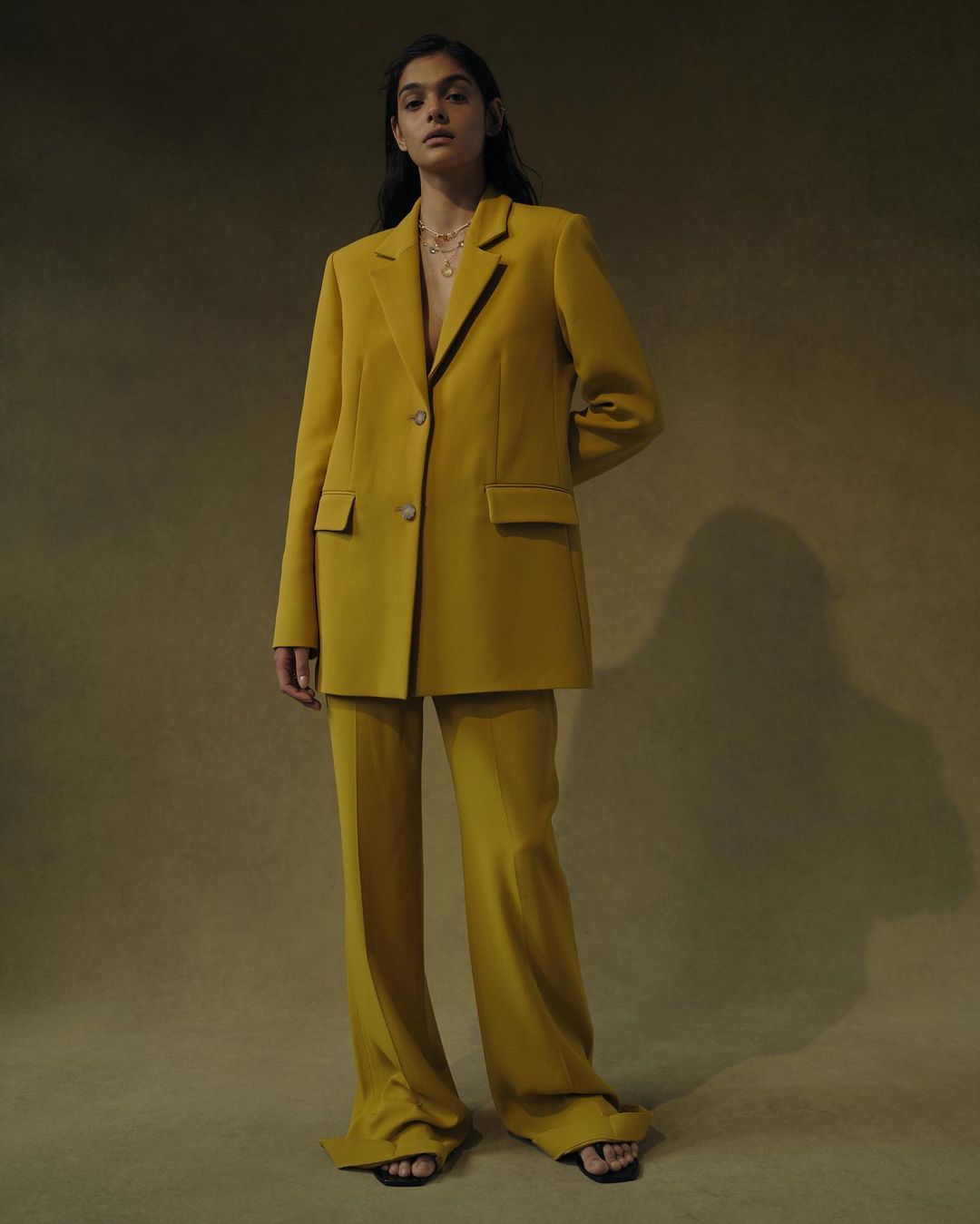 Woman poses in a yellow oversized blazer and matching pants on tan backdrop in a studio setting, fashion shoot, looking at the camera.