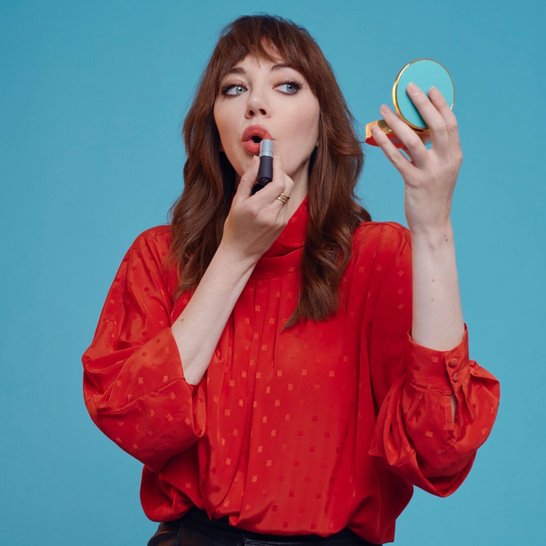 Brunette woman with red top applies lipstick while holding a compact mirror against a bright blue backdrop in a medium shot.
