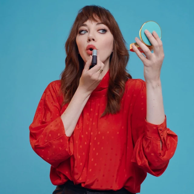Brunette woman with red top applies lipstick while holding a compact mirror against a bright blue backdrop in a medium shot.