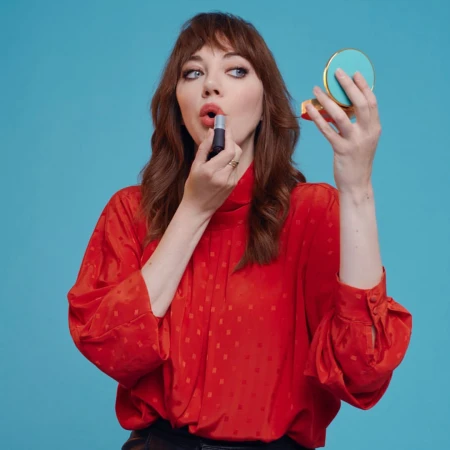 Brunette woman with red top applies lipstick while holding a compact mirror against a bright blue backdrop in a medium shot.