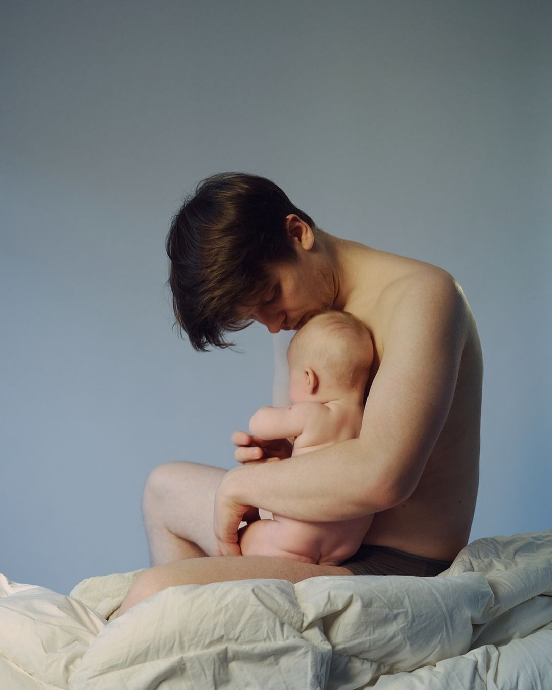 A fair-skinned adult male sits on a bed, cradling a fair-skinned baby with a gray background. The subjects are in a soft, diffused light.