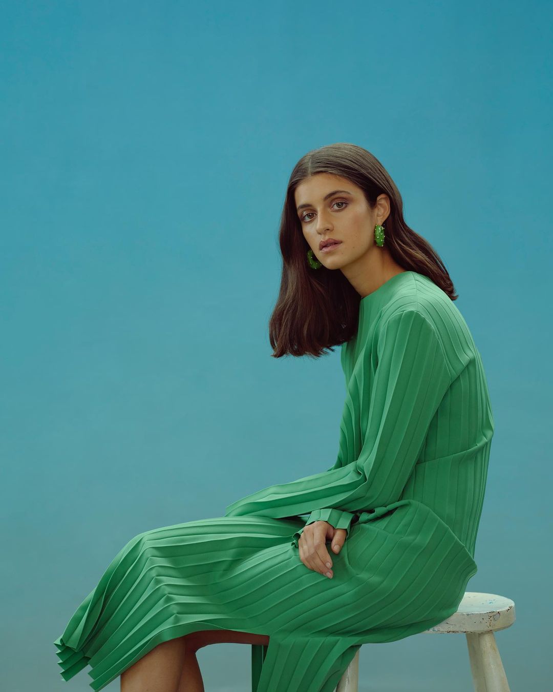A brunette woman in a green pleated dress sits on a stool against a teal background, gazing at the camera with a calm expression.