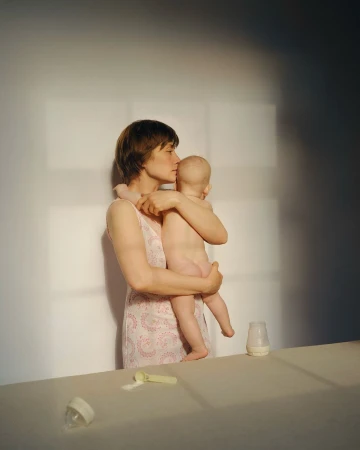 Indoor portrait of a mother gently kissing her baby's head in a warm, soft light, with baby bottles and formula on the table nearby.