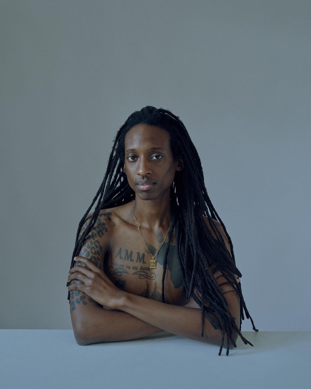 Portrait of a slender Black adult with long dreadlocks, tattoos, and a gold necklace, posing indoors with arms crossed on a white surface.