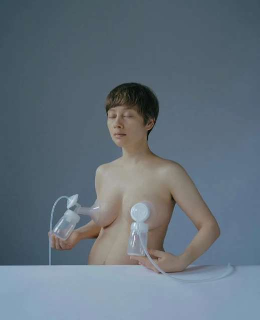 Nude woman with breast pump, looking down in a softly lit gray room, holding milk collection bottles at a white table.