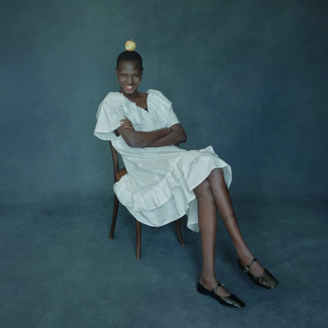 Young Black woman in a white dress sitting in a chair with an apple on her head, studio lighting against a dark gray backdrop.