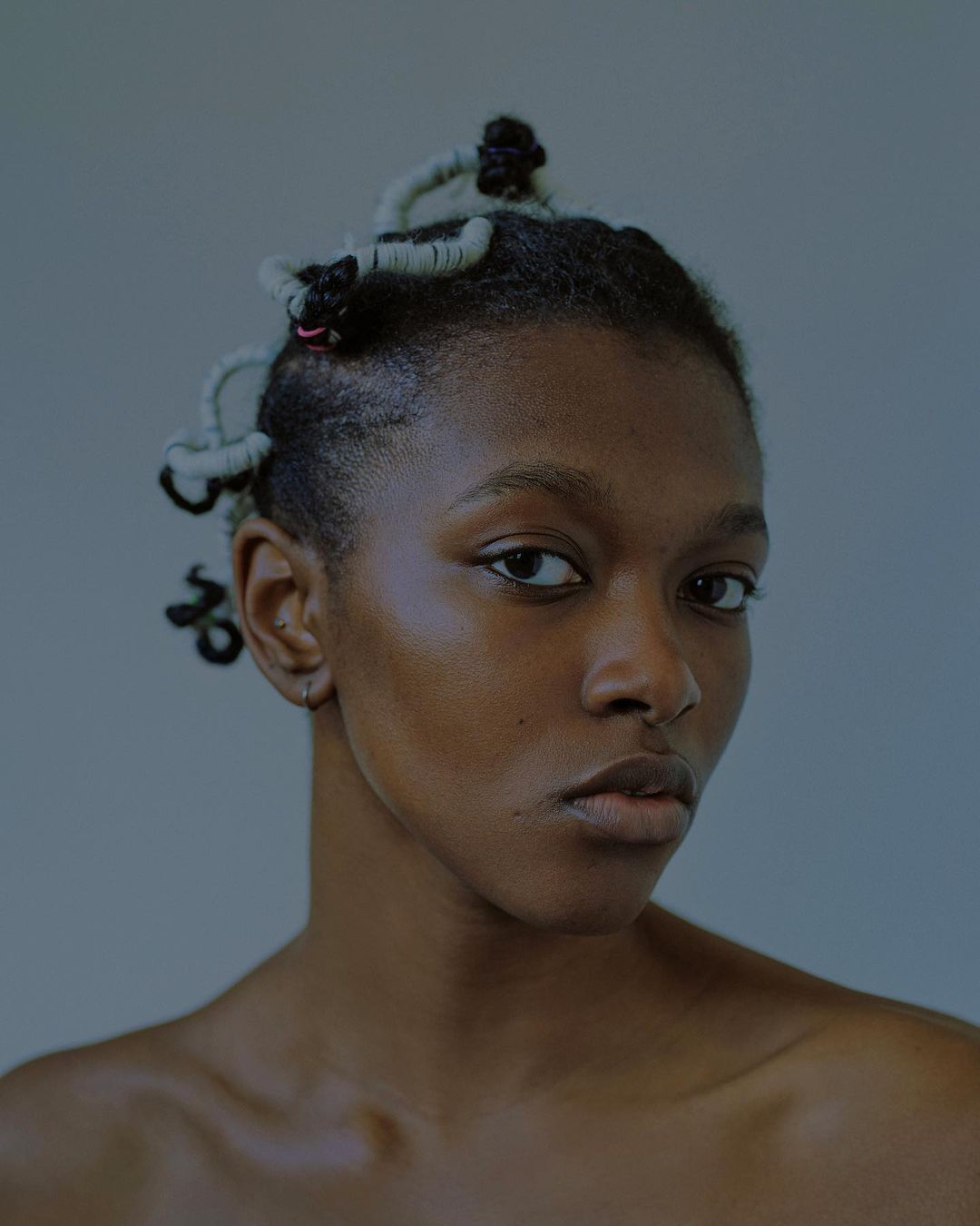 Close-up portrait of a young African woman with an expressive gaze, unique twisted hairstyle against a solid, muted blue-grey background.