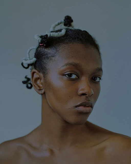 Close-up portrait of a young African woman with an expressive gaze, unique twisted hairstyle against a solid, muted blue-grey background.