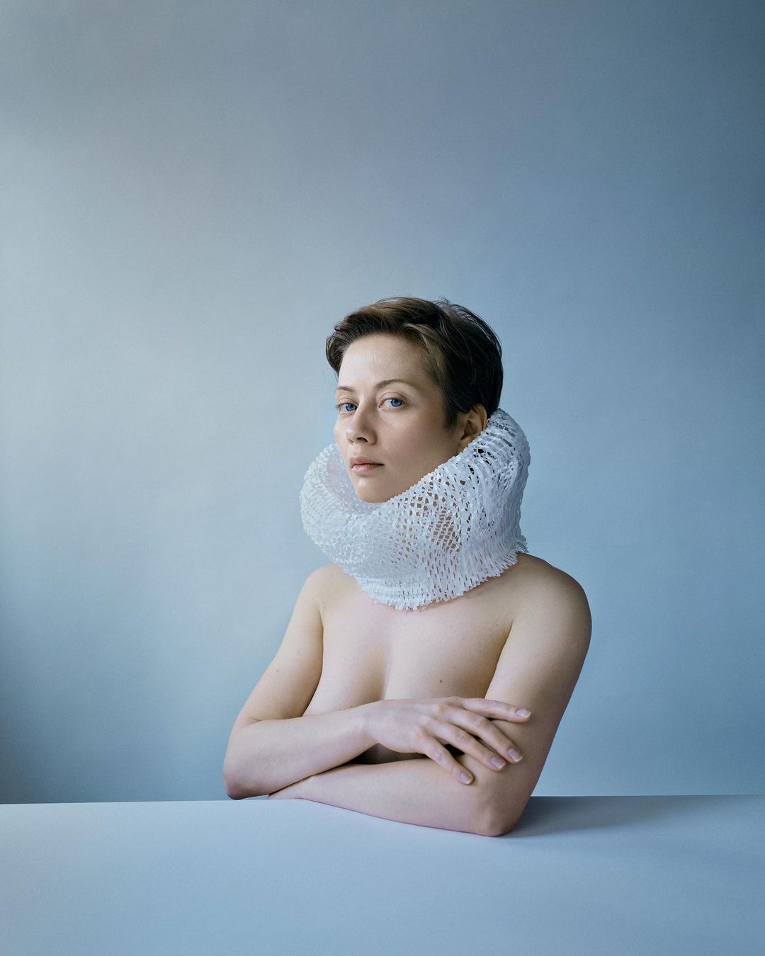 Portrait of a fair-skinned adult woman with short brown hair, wearing a white net collar, against a pale blue background and white tabletop.
