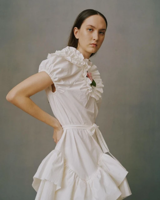 Portrait of a woman in a white ruffle dress with hands on her hips, standing against a plain gray backdrop in a fashion editorial style.