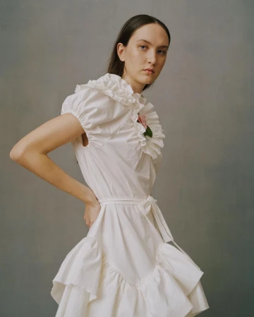 Portrait of a woman in a white ruffle dress with hands on her hips, standing against a plain gray backdrop in a fashion editorial style.