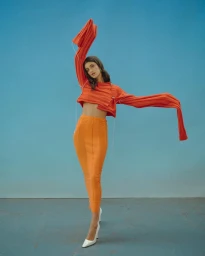Model in orange cropped sweater and matching skirt posing against a blue studio background