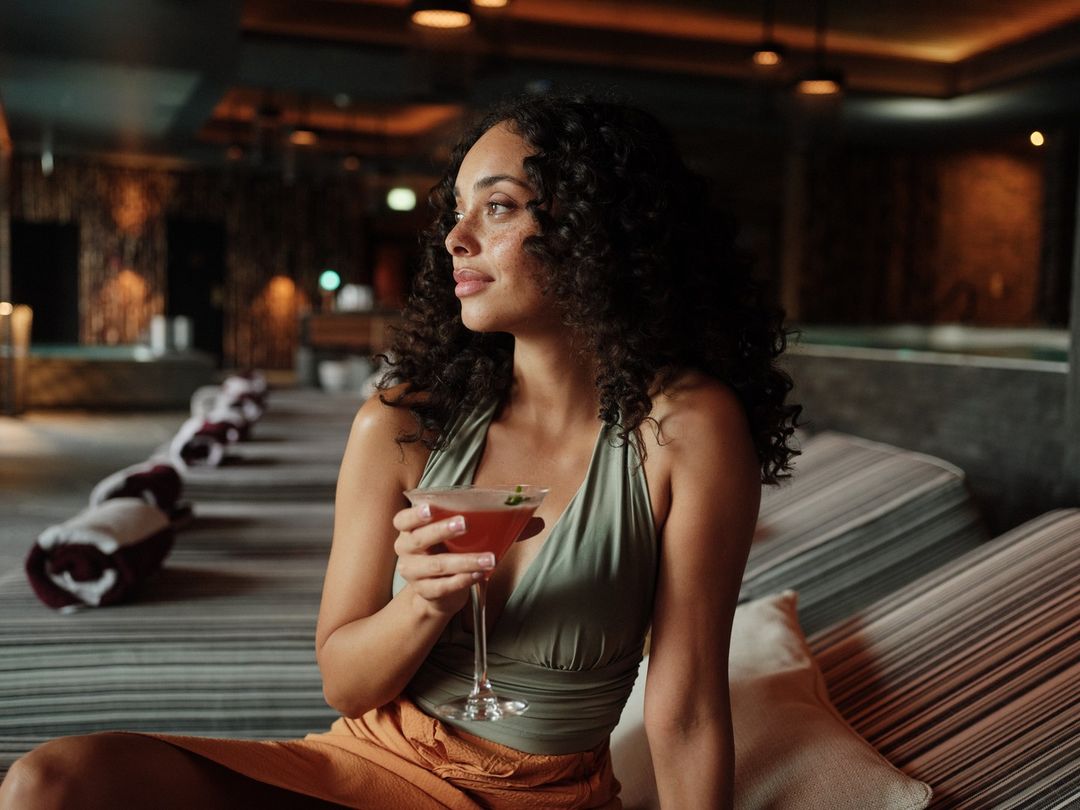 Woman in a spa setting lounges with a cocktail. She is surrounded by relaxing lounge beds and soothing warm lighting.