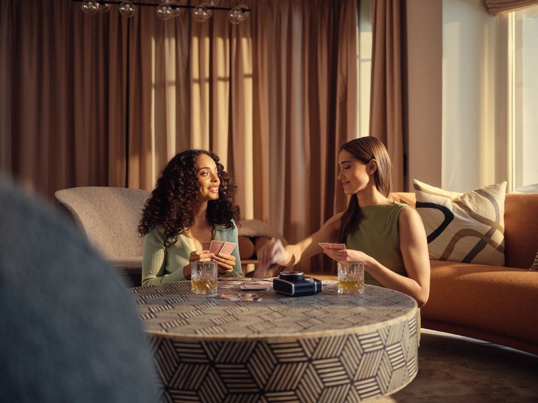 Two women are sitting at a round table playing cards with drinks in a warm, inviting room with neutral colors and soft, natural lighting.
