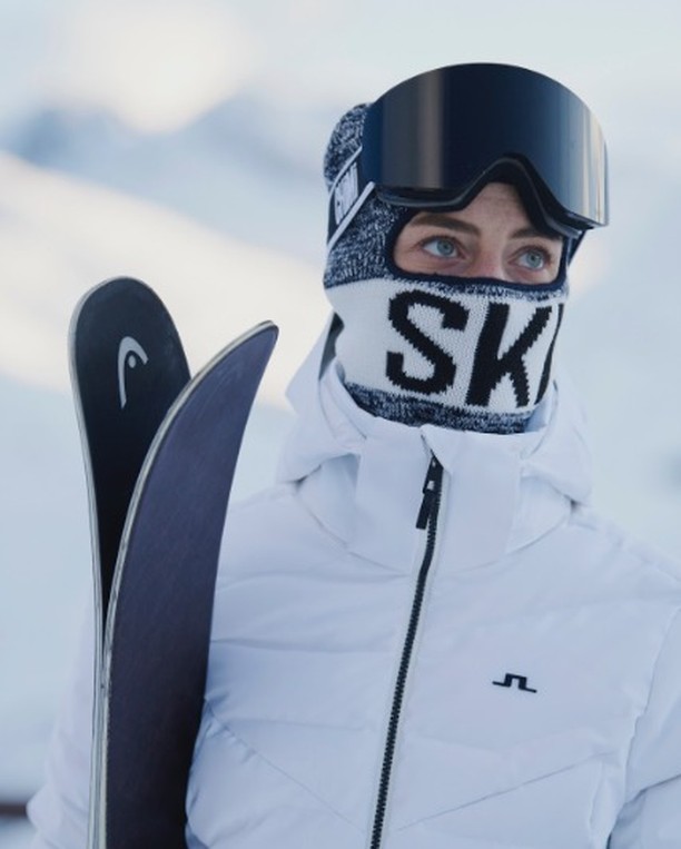Person wearing ski goggles and a ski mask that reads "SKI" in the snow, holding skis. The person is wearing a white jacket.