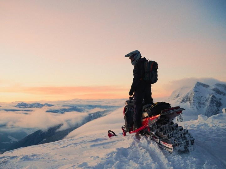 Person in helmet and backpack stands next to snowmobile on snowy mountain peak overlooking clouds and mountain range at sunset.