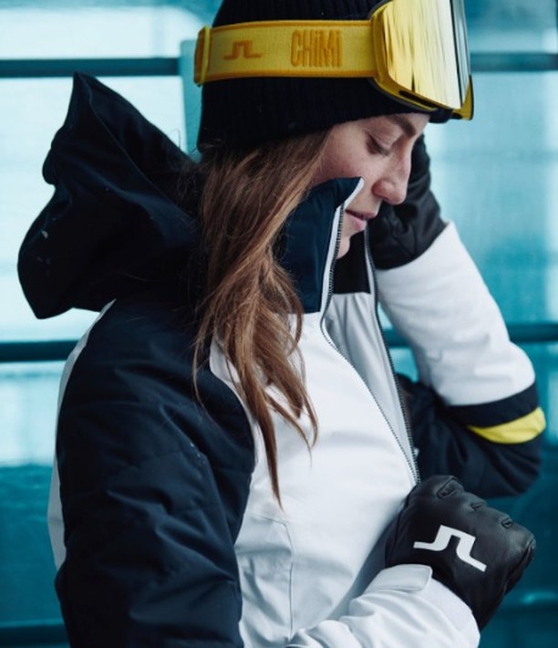 Woman in black beanie and ski goggles zips up a white ski jacket with black accents, wearing black gloves with a white logo.