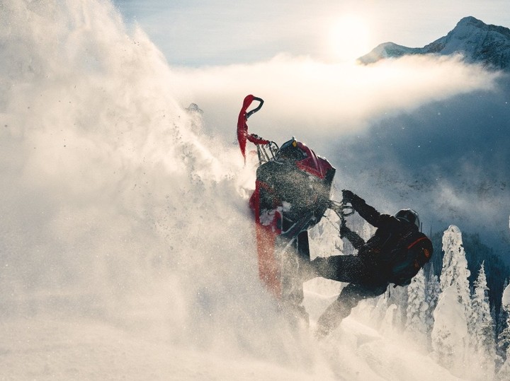 Snowmobiler performing aerial stunt, covered in snow, with mountain backdrop, sunlit sky, and evergreens dusted with snow.