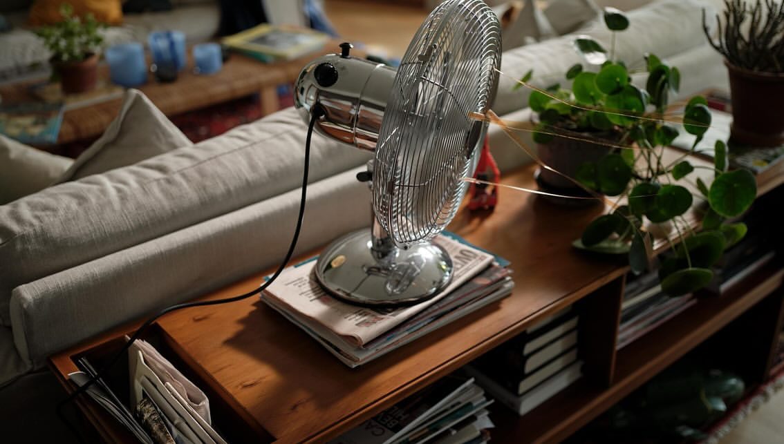 Indoor shot of a silver vintage electric fan placed on a wooden shelf, with a stack of newspapers, a potted plant, and part of a couch visible.