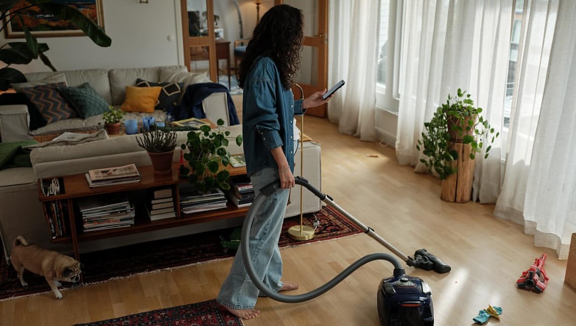 Adult female vacuums a light wood floor in a brightly lit living room with large windows and sheer white curtains, with a dog nearby.