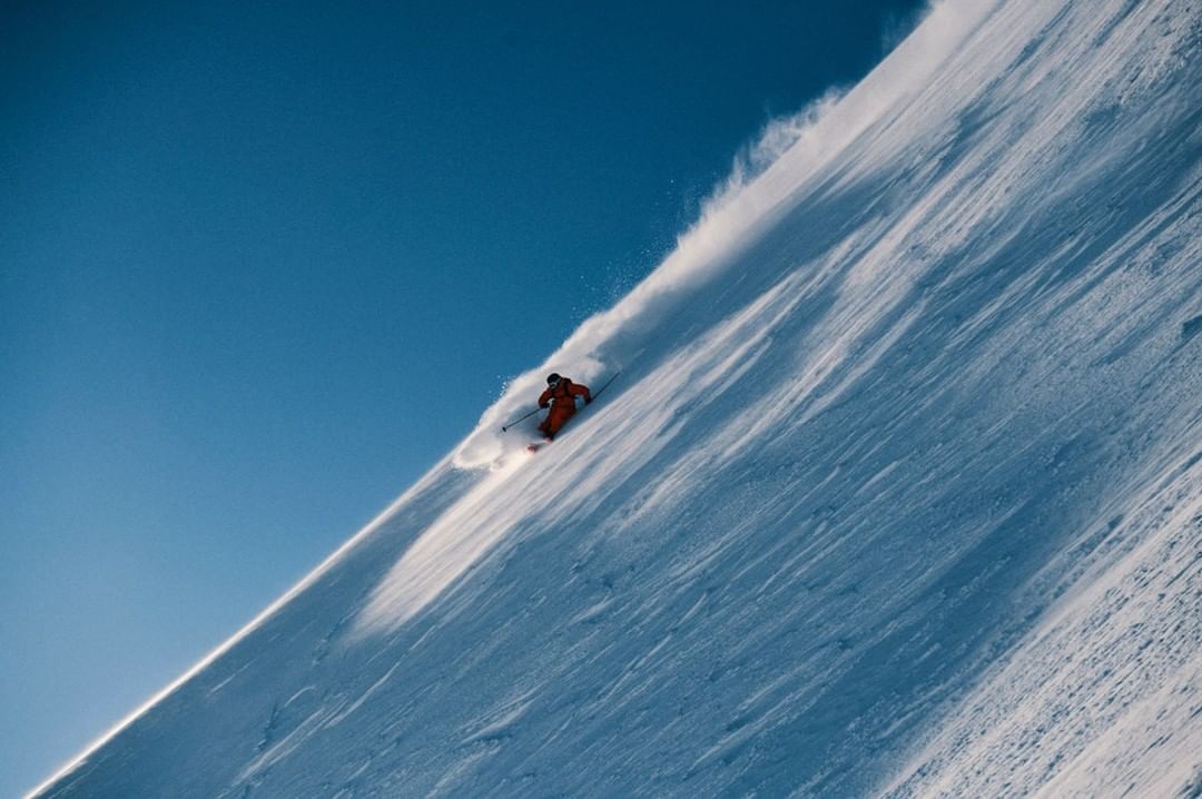 Person in orange ski suit skiing down a steep snow-covered mountain on a bright sunny day with a clear blue sky.